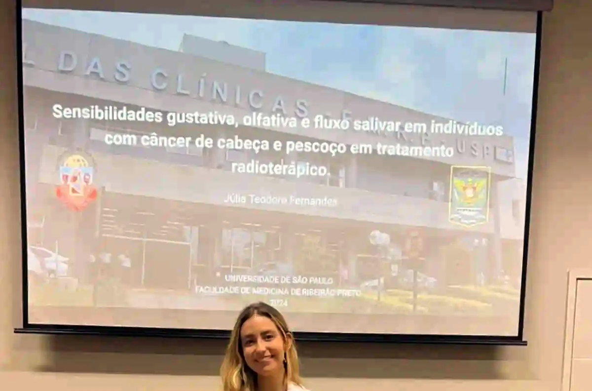 Júlia diante do telão da defesa do mestrado, com o título da dissertação projetado e os brasões da USP e da Faculdade de Medicina de Ribeirão Preto