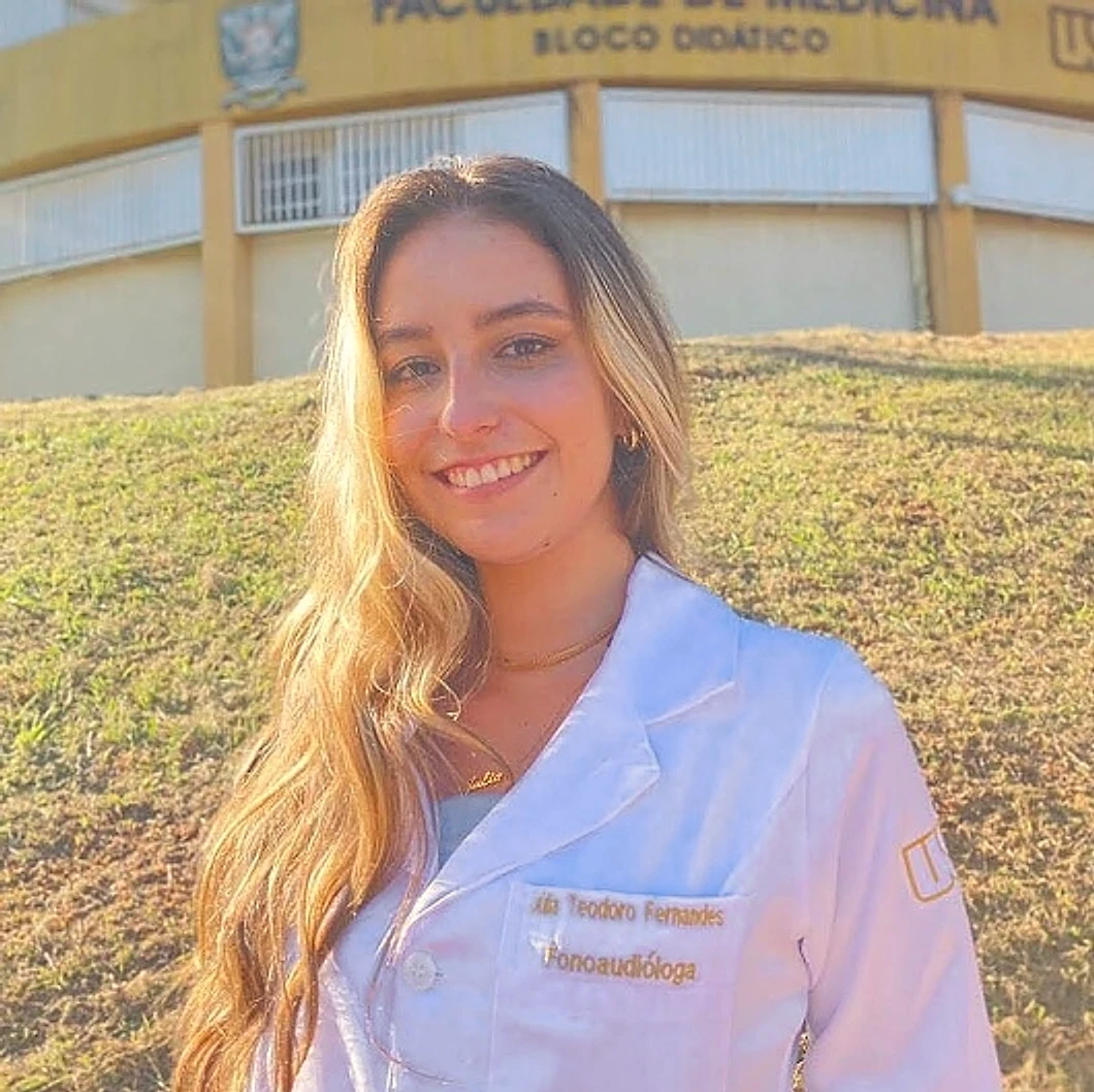 Júlia Teodoro sorrindo, de jaleco branco com o nome bordado, diante do Bloco Didático da Faculdade de Medicina de Ribeirão Preto, numa tarde de sol